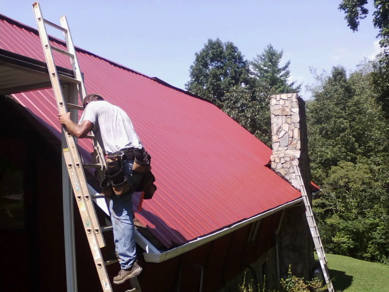 Roofer on a ladder beside a red metal roof and stone chimney — Soffit & Fascia Repair by Roofing Brothers in Linesville